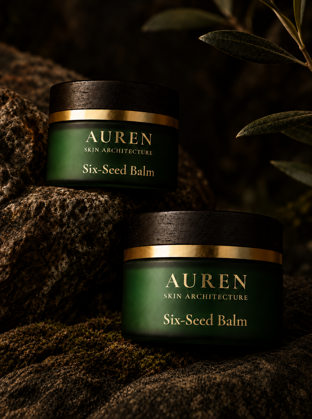 Two jars of Auren Skin Architecture Six-Seed Balm arranged on mossy rocks with olive leaves in the background. The jars are dark green with black lids and gold bands, set against a dark, natural outdoor backdrop.