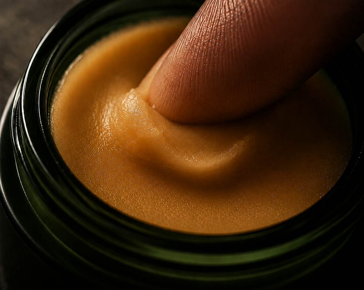 A close-up shot of a finger dipping into a thick, smooth, golden-orange balm or cream inside a dark green glass jar.